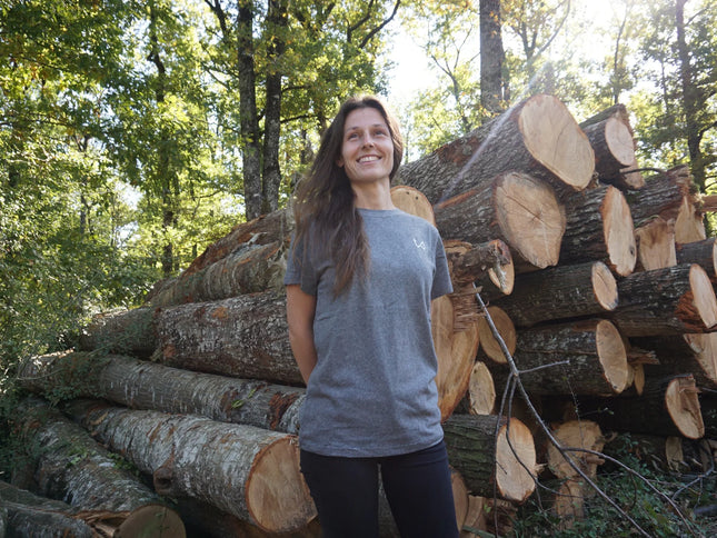Woman standing next to a stack of logs in a forest wearing hemp and organic cotton tshirt.