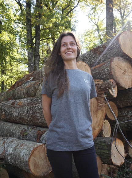 Woman standing next to a stack of logs in a forest wearing hemp and organic cotton tshirt.