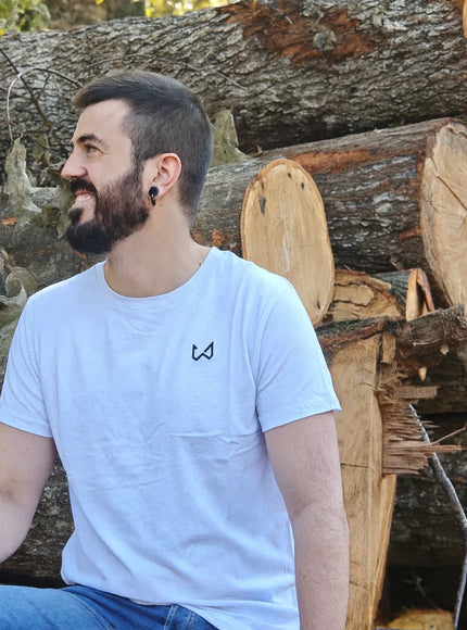 Man sitting among stacked logs wearing a white t-shirt with a logo of wild dynamics