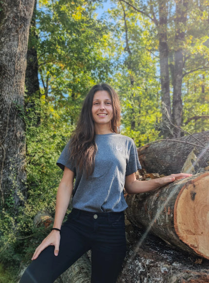 Woman standing next to a large log in a forest wearing gray organic cotton tshirt of wild dynamics