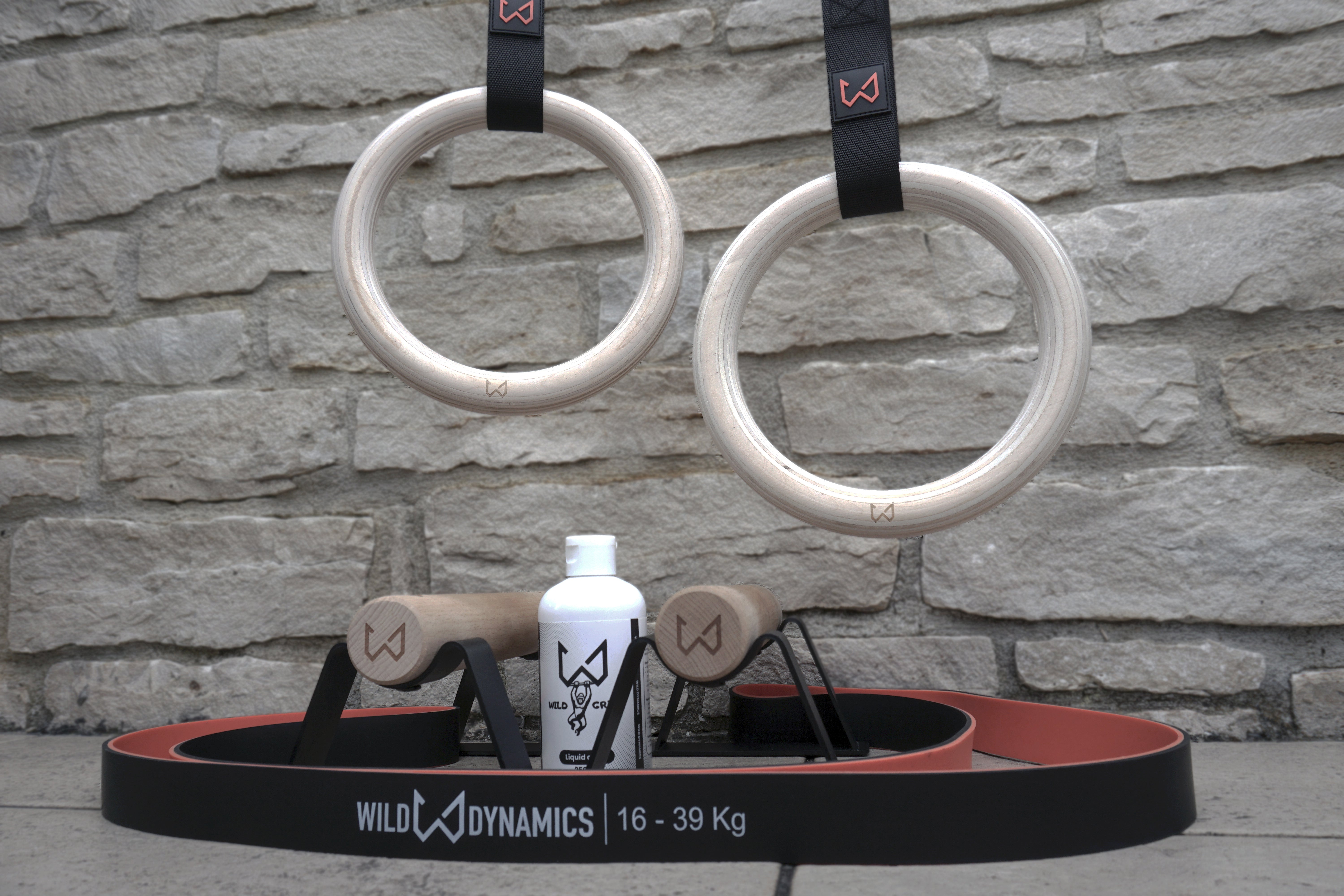 Calisthenics equipment set, wooden gymnastic rings, resistance band, wooden parallettes, liquid chalk, displayed against a stone wall background.