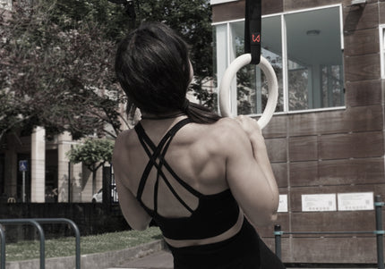 A female calisthenics athlete with muscular back performing a pull-up on workout wooden rings outdoors with a building in the background