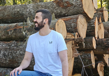 Man sitting among stacked logs wearing a white t-shirt with a logo of wild dynamics