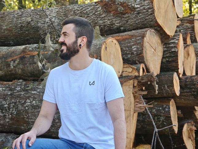 Man sitting among stacked logs wearing a white t-shirt with a logo of wild dynamics
