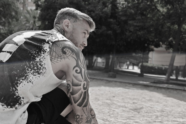 Athlete with tattoos on his arms wearing white tank top in a calisthenics park with trees in the background