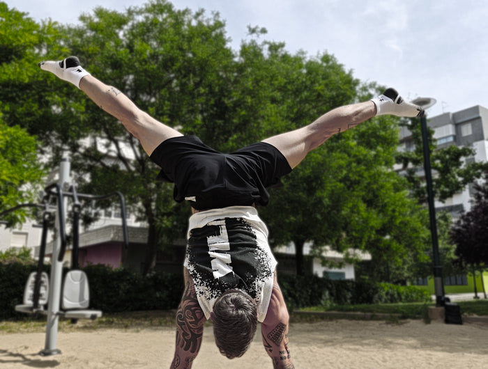 Athlete doing a handstand wearing a white tank top made from hemp on small parallettes by wild dynamics with trees and buildings in the background