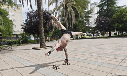 Tattooed calisthenics athlete performing a one-arm handstand on small parallettes, with palm trees and buildings in the background.