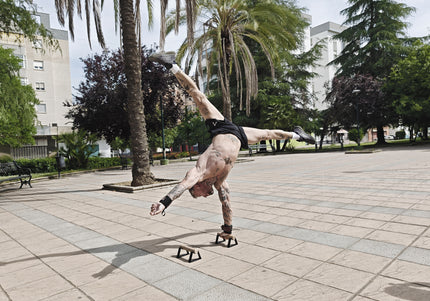Tattooed calisthenics athlete performing a one-arm handstand on small parallettes, with palm trees and buildings in the background.