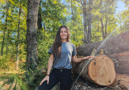 Woman standing next to a large log in a forest wearing gray organic cotton tshirt of wild dynamics