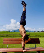 Person doing a handstand on a wooden bench in an open field with mountains in the background.