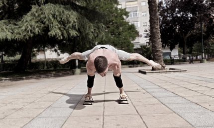 Calisthenics athlete performing a planche on small parallettes wearing elbow sleeves in an urban park setting.