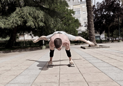 Calisthenics athlete performing a planche on small parallettes wearing elbow sleeves in an urban park setting.