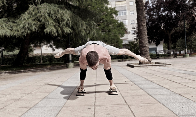 Calisthenics athlete performing a planche on small parallettes wearing elbow sleeves in an urban park setting.