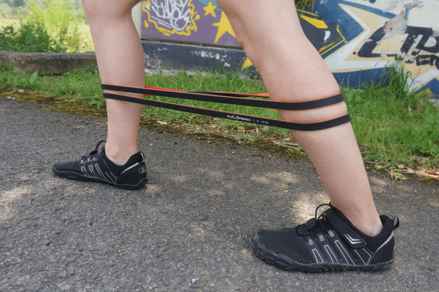 Person using resistance bands outdoors on a paved path with grass and a colorful mural in the background.