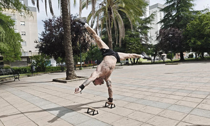 Person performing a one arm handstand on parallettes in a park.